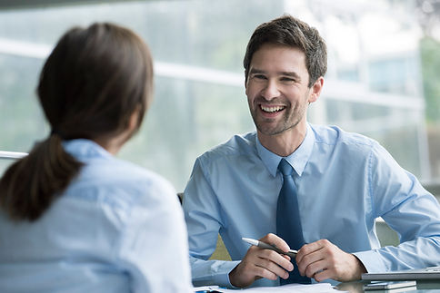 A professional man in a light blue shirt and tie smiles while engaging in conversation with a woman during a meeting.