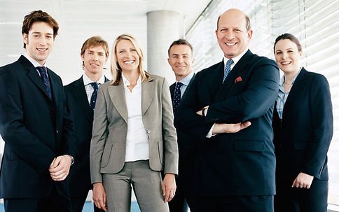 A group of six professionals, dressed in business attire, smiling and posing together in a modern office environment with large windows.