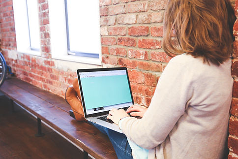 A person sitting on a wooden bench working on a laptop in a brick-walled environment.